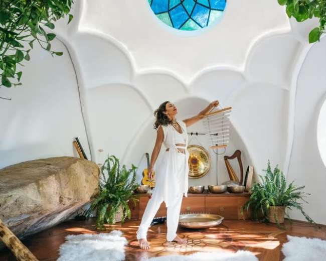 A woman in white clothing stands in a brightly lit, circular room with natural elements and musical instruments.