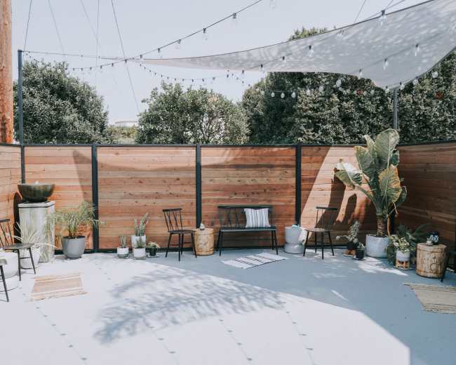 The image shows a modern outdoor space with wooden fencing, string lights overhead, and various seating arrangements surrounded by potted plants.