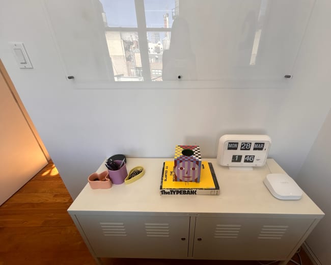A white storage unit displays various items, including a clock, a lamp, books, and pencil holders, with a window in the background letting in natural light.