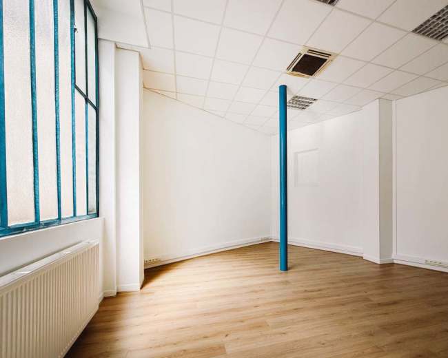 The image shows an empty room with wooden flooring, a large window, and a blue pillar in the center.