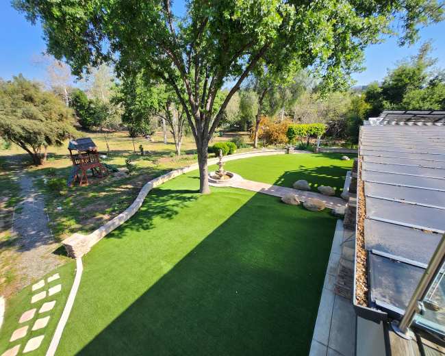 The image shows a landscaped yard with artificial grass, a stone pathway, and a large tree, surrounded by various shrubs and a wooden play structure in the background.