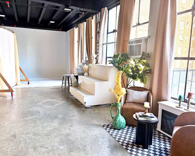 The image shows a spacious studio with large windows, featuring a seating area, decorative plants, and a white backdrop set for photography.