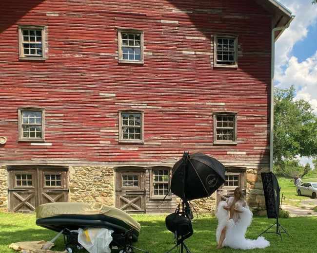 A woman in a white dress poses near photography equipment in front of a red barn with multiple windows.