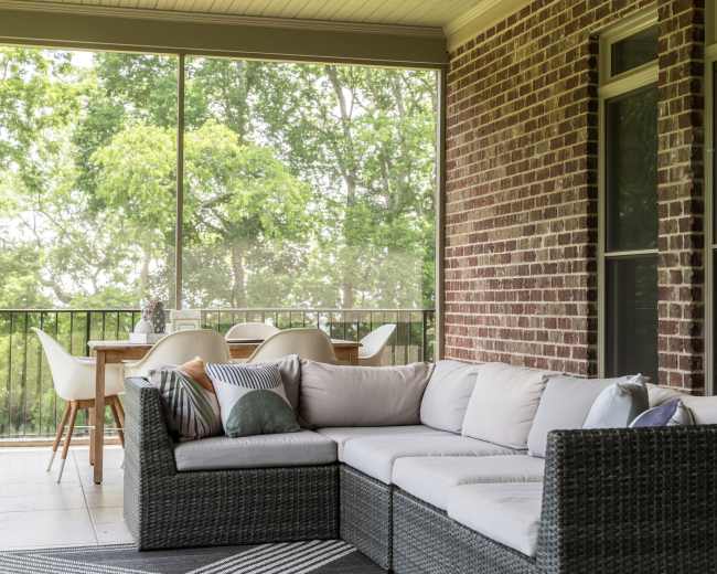 A screened patio features a sectional sofa and a dining table with chairs, surrounded by greenery.