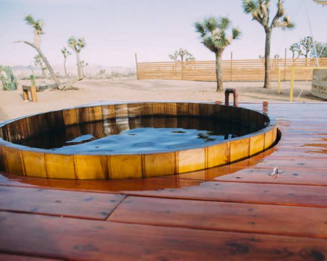 A circular wooden hot tub is set on a red wooden deck in a desert landscape, with Joshua trees in the background.