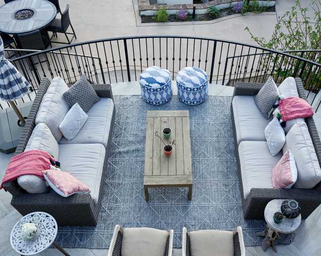 A outdoor seating area features a central coffee table surrounded by gray couches, patterned poufs, and chairs, overlooking a swimming pool and landscaped garden.