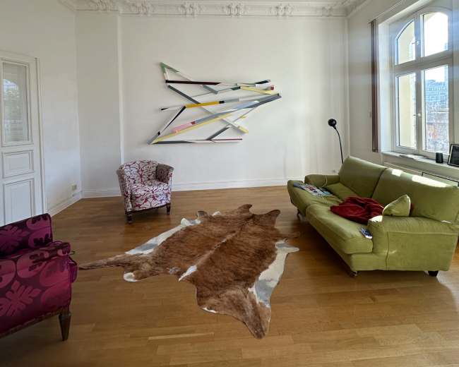 The image shows a spacious living room featuring a green sofa, a patterned armchair, a cowhide rug on the wooden floor, and abstract wall art above.