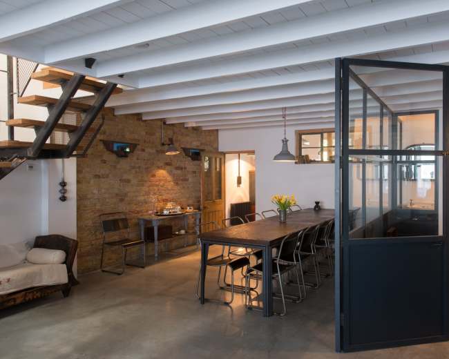 The image shows a modern, open-concept space featuring a dining area with a long table and chairs, a staircase to the left, and glass partitions separating different sections.