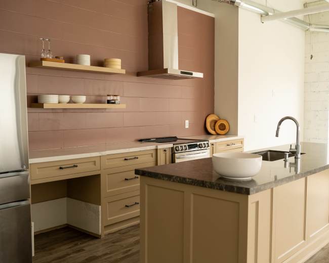 The image shows a modern kitchen with beige cabinetry, a dark countertop, and a wall with a reddish-brown panel.