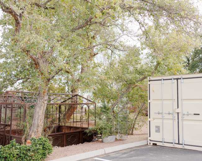 A large shipping container is displayed next to a metal greenhouse surrounded by trees in a parking area.
