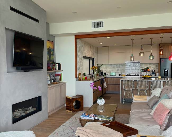 The image shows a modern living room with a fireplace and a view of an open kitchen, featuring a mix of light wood and gray decor.