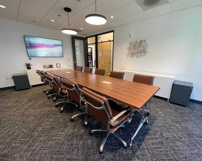 A large conference room features a long wooden table with eight rolling chairs, a television on the wall, and two filing cabinets in the corner.