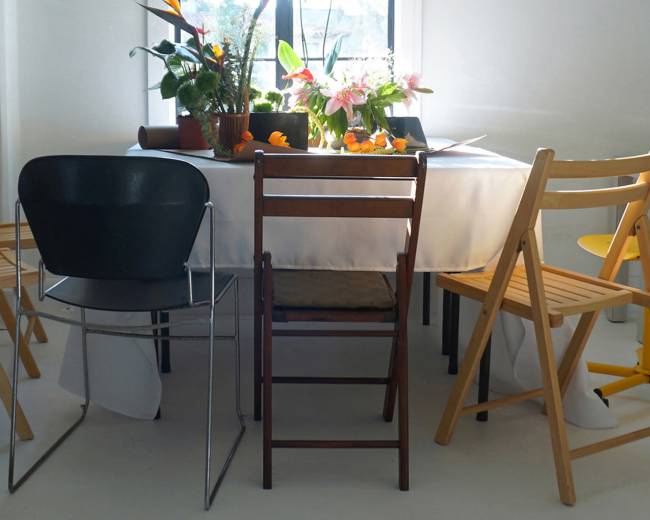 A round table with a white tablecloth is surrounded by various types of chairs, and there are potted plants and flowers on the table.