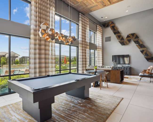 The image shows a modern lounge area with large windows, a pool table, stylish furnishings, and a view of an outdoor pool area.