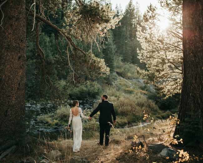 A bride and groom walk hand in hand along a forested path beside a stream, with sunlight filtering through the trees.