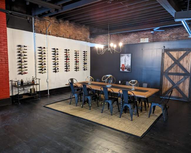 A dining area with a long wooden table surrounded by black metal chairs, a wine display on the wall, and a sliding barn door in a rustic setting.