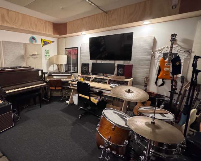 The image shows a small music studio featuring various instruments, including a drum set, a piano, and several guitars, along with sound equipment and storage shelves.