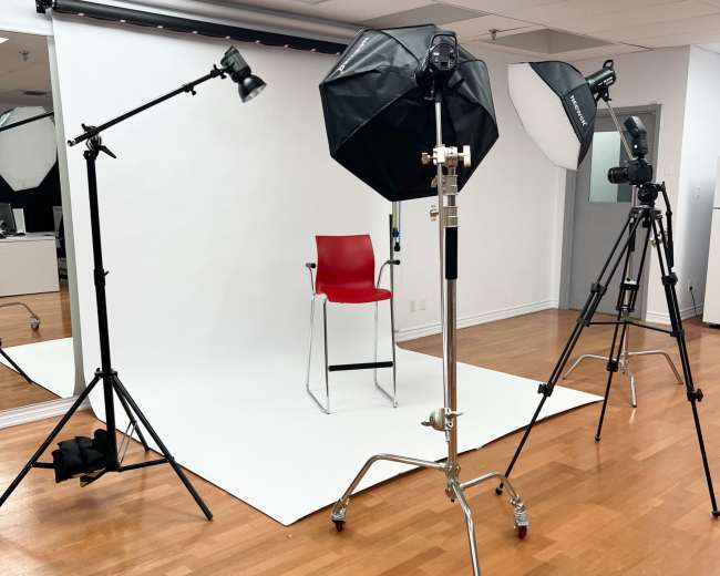 The image shows a photography studio setup with lighting equipment, a camera on a tripod, and a red chair on a white backdrop.