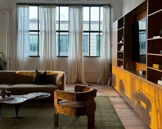 A modern living room features large windows with sheer curtains, a brown sofa, a wooden cabinet, and a brown accent chair on a green area rug.