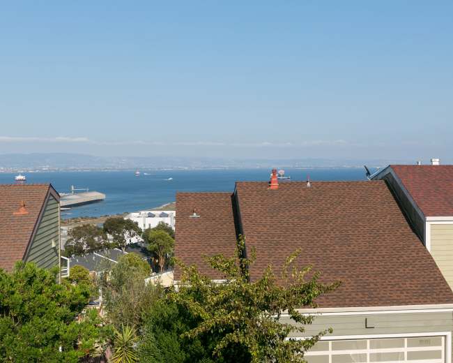The image shows a coastal view from a residential area, with rooftops in the foreground and a blue ocean in the background, dotted with ships.