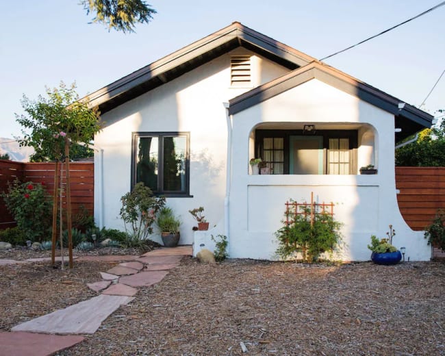 A white cottage-style house with a paved pathway leads to a front porch, surrounded by a garden and wooden fence.