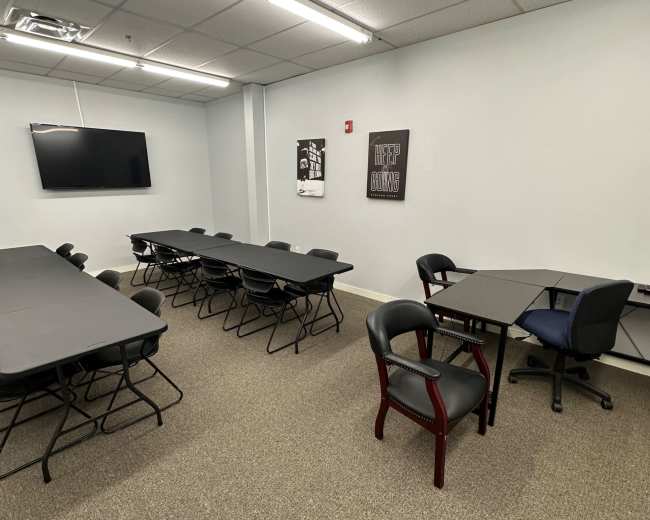 A conference room with several folding tables and chairs arranged in an open layout, along with a wall-mounted TV and framed posters on the walls.