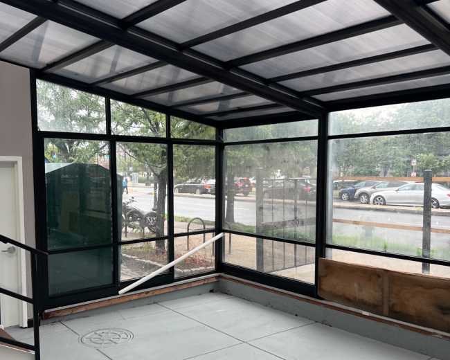 The image shows a glass-enclosed room with concrete flooring and visible rain on the windows, overlooking a street with parked cars and trees outside.