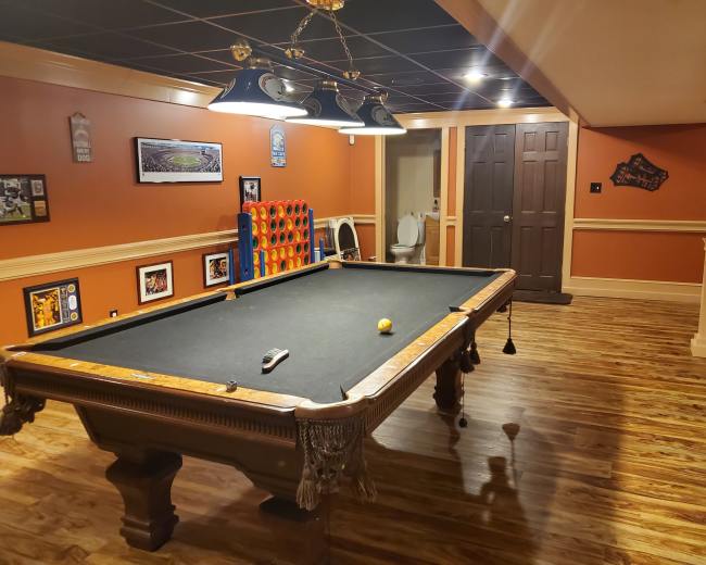The image shows a recreation room featuring a pool table, wooden flooring, orange walls, framed pictures, and a bathroom door in the background.