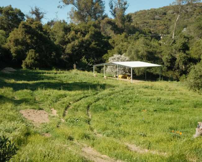 A grassy area features a covered picnic structure surrounded by trees and tire tracks leading into the field.