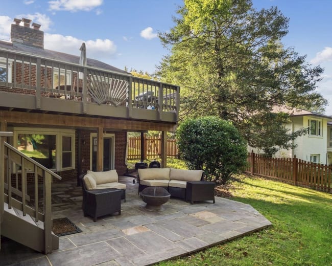 The image shows a backyard with a patio featuring a seating area, a fire pit, and a deck above surrounded by trees and a fenced yard.