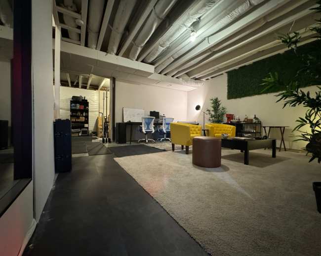 The image depicts a modern workspace featuring two blue chairs at a desk, a yellow couch, a round brown table, and green plants, all situated in a room with exposed ceiling beams and a carpeted floor.