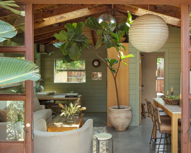 The image shows a modern, open-concept living space with a wooden ceiling, large windows, a dining area, and a potted plant.