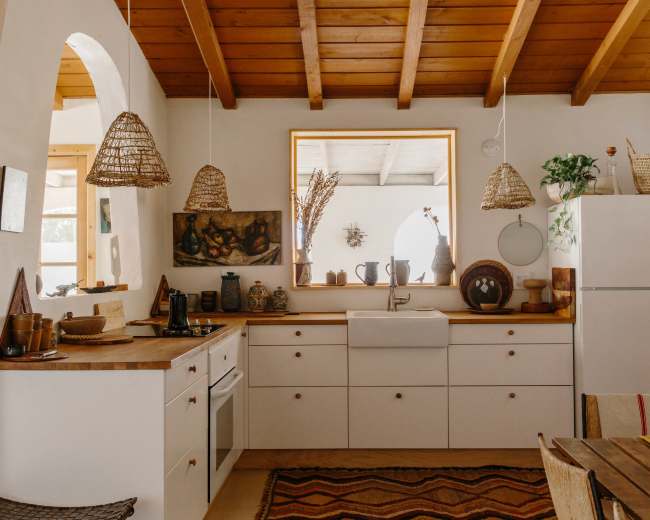The image shows a cozy kitchen with wooden beams, white cabinetry, a farmhouse sink, and woven pendant lights.