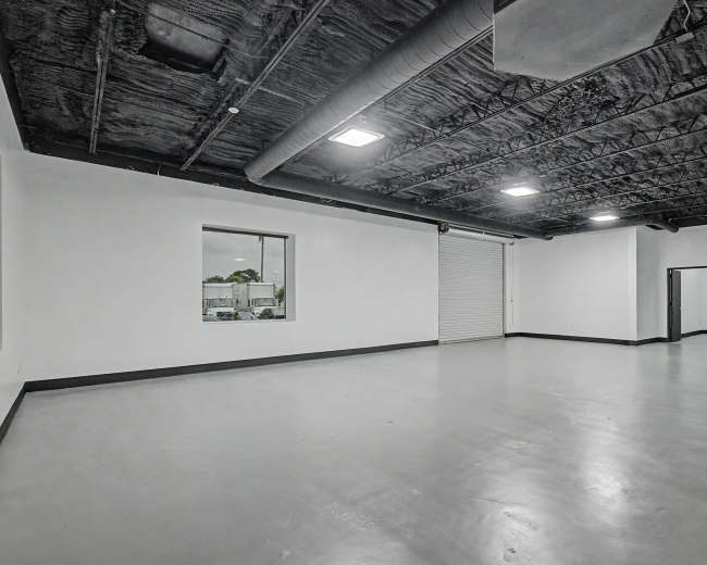 The image shows an empty room with white walls, a concrete floor, and a black ceiling, featuring windows and a roll-up door.