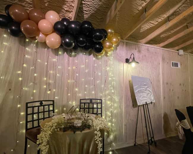 A decorated indoor space featuring a table with a geometric centerpiece, a backdrop of balloons in black, gold, and brown, fairy lights, and a framed sign on an easel.