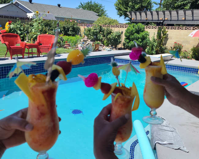 Three colorful cocktails are raised in front of a swimming pool surrounded by greenery and lounge chairs.