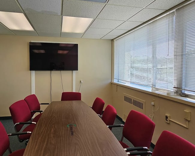 A conference room features a long wooden table surrounded by red chairs, with a wall-mounted TV and large windows covered by blinds.