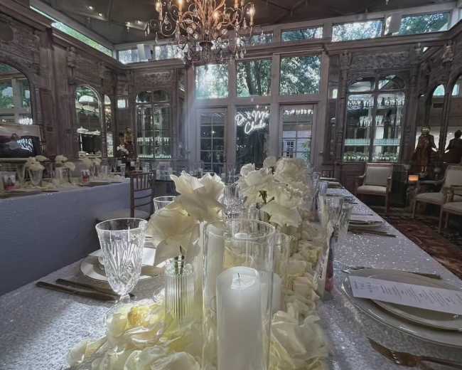 An elegantly set dining table adorned with white flowers and candles, surrounded by a lavishly decorated interior featuring large windows and a chandelier.
