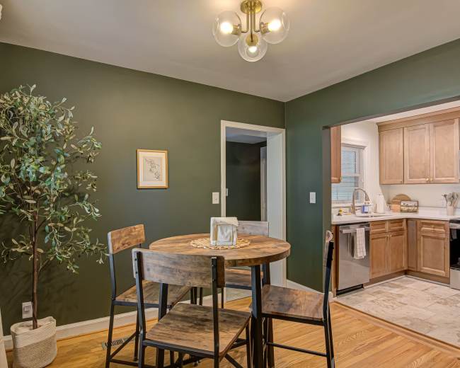 The image shows a dining area featuring a round wooden table with four chairs, a potted plant, and an open kitchen visible through a doorway.