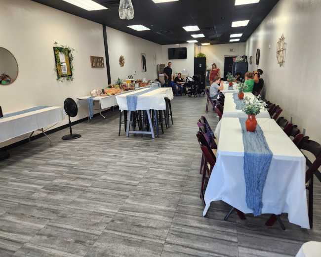 The image shows a spacious interior of a dining area with tables set for an event, some food displayed on a side table, and a small group of people at the back.