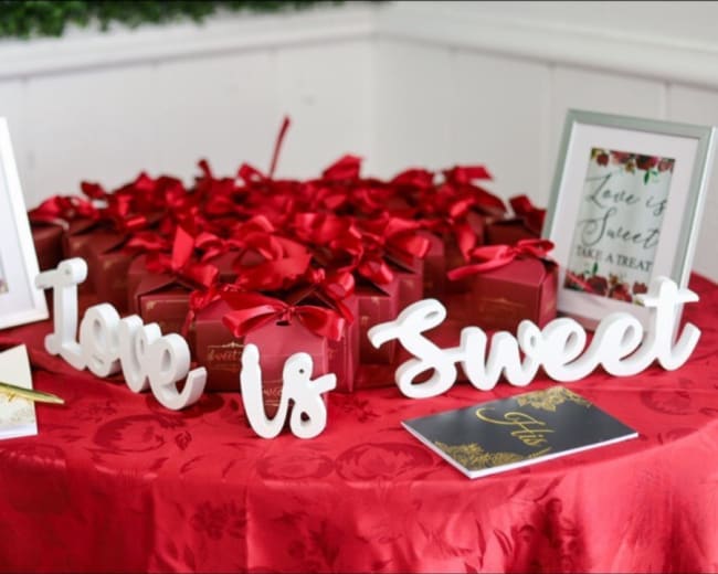 A table is decorated with a red tablecloth, featuring gift boxes tied with ribbons, framed signs, and a decorative message that reads "Love Is Sweet."