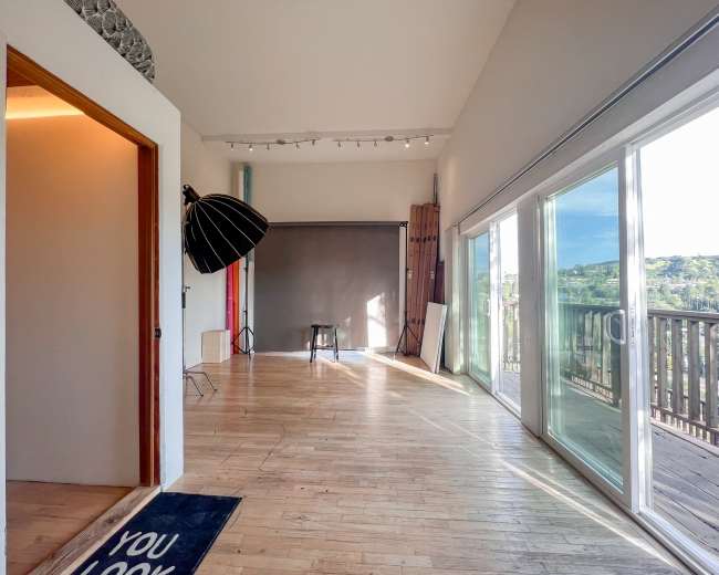 The image shows a spacious, well-lit room with large windows, a wooden floor, and photography equipment, including a large umbrella light and a stool.