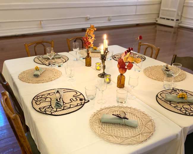 A set dining table is arranged with woven placemats, glassware, and small flower arrangements, illuminated by candles in brown glass vases.