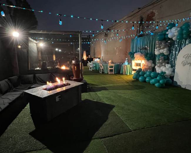 An outdoor party setup featuring a fire pit, a seating area with a dark sofa, tables decorated with teal balloons, and ambient lighting.