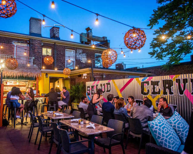 A bustling outdoor bar scene features patrons seated at tables under string lights, with a vibrant mural and a food counter visible in the background.