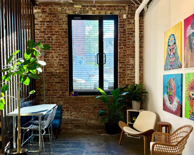 The image shows a stylish room with exposed brick walls, a window, artwork on the wall, and a mix of modern furniture including chairs and a table.