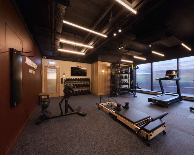 The image shows a modern gym interior with various fitness equipment, including weights, a treadmill, and a reformer, set against a backdrop of large windows.