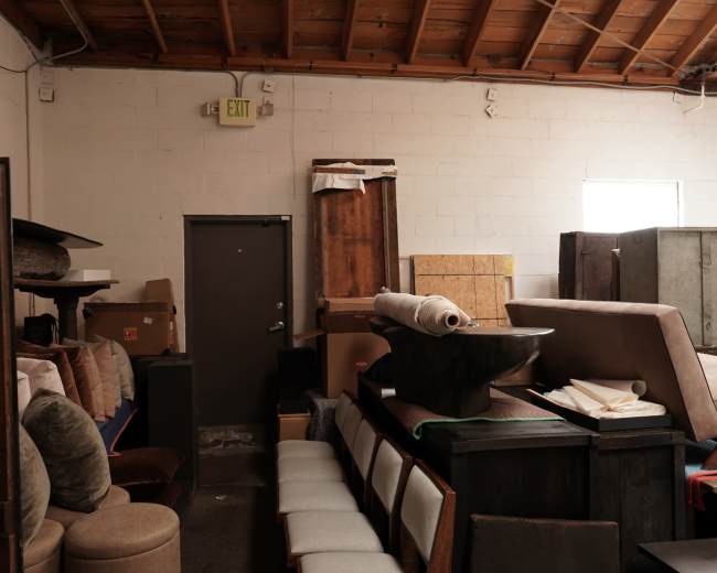 The image shows a cluttered warehouse space filled with various pieces of furniture and boxes stacked against the walls.