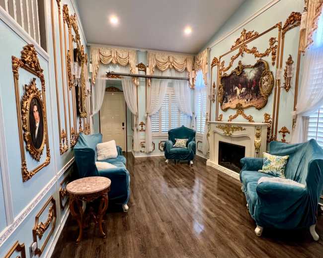 The room features blue walls adorned with decorative molding and framed portraits, along with two turquoise armchairs and a fireplace.