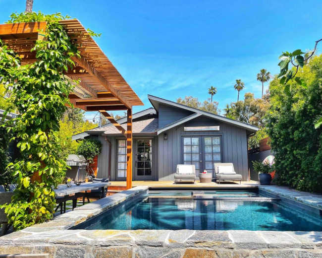 A modern house with a gray exterior sits beside a swimming pool surrounded by greenery and patio furniture.